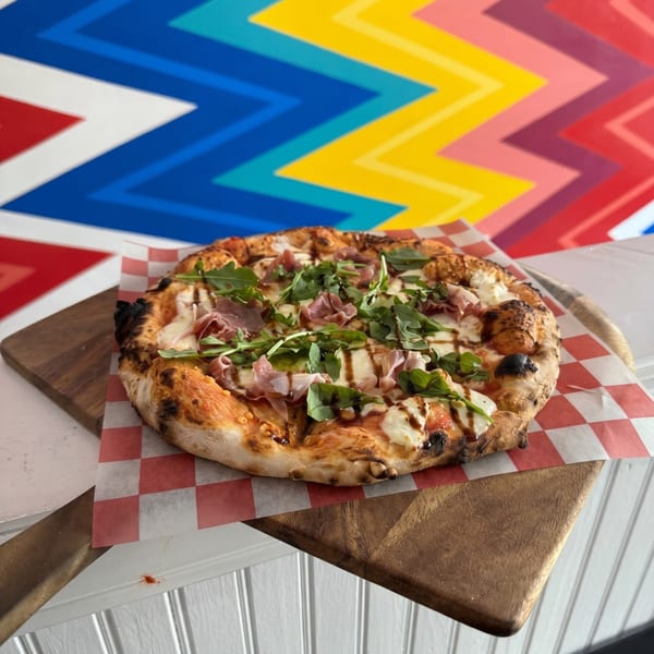 A pizza with prosciutto, arugula, and balsamic glaze on a wooden board with a colorful background.
