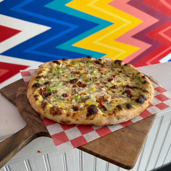 A freshly baked pizza with pesto, cheese, and vegetables on a wooden peel against a colorful wall.