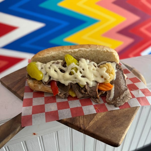 A beef sandwich with peppers and white sauce on a wooden board with a colorful background.