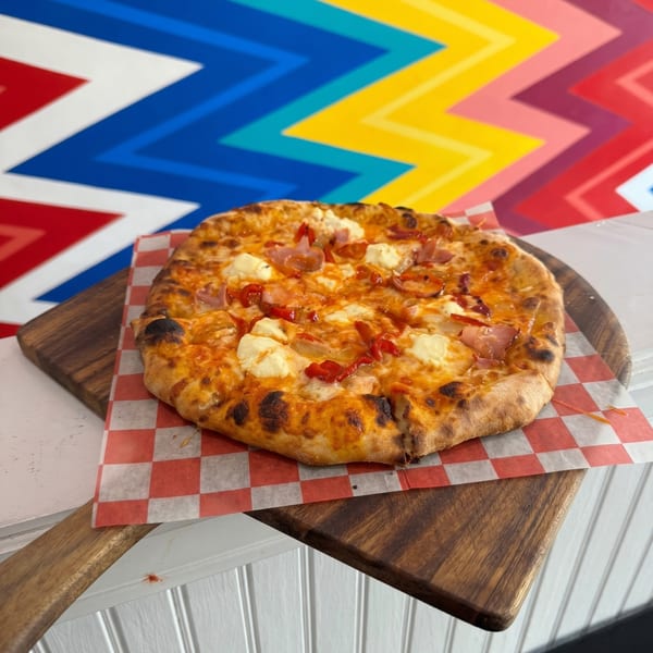 A freshly baked pizza with cheese, red peppers, and ham on a wooden peel.